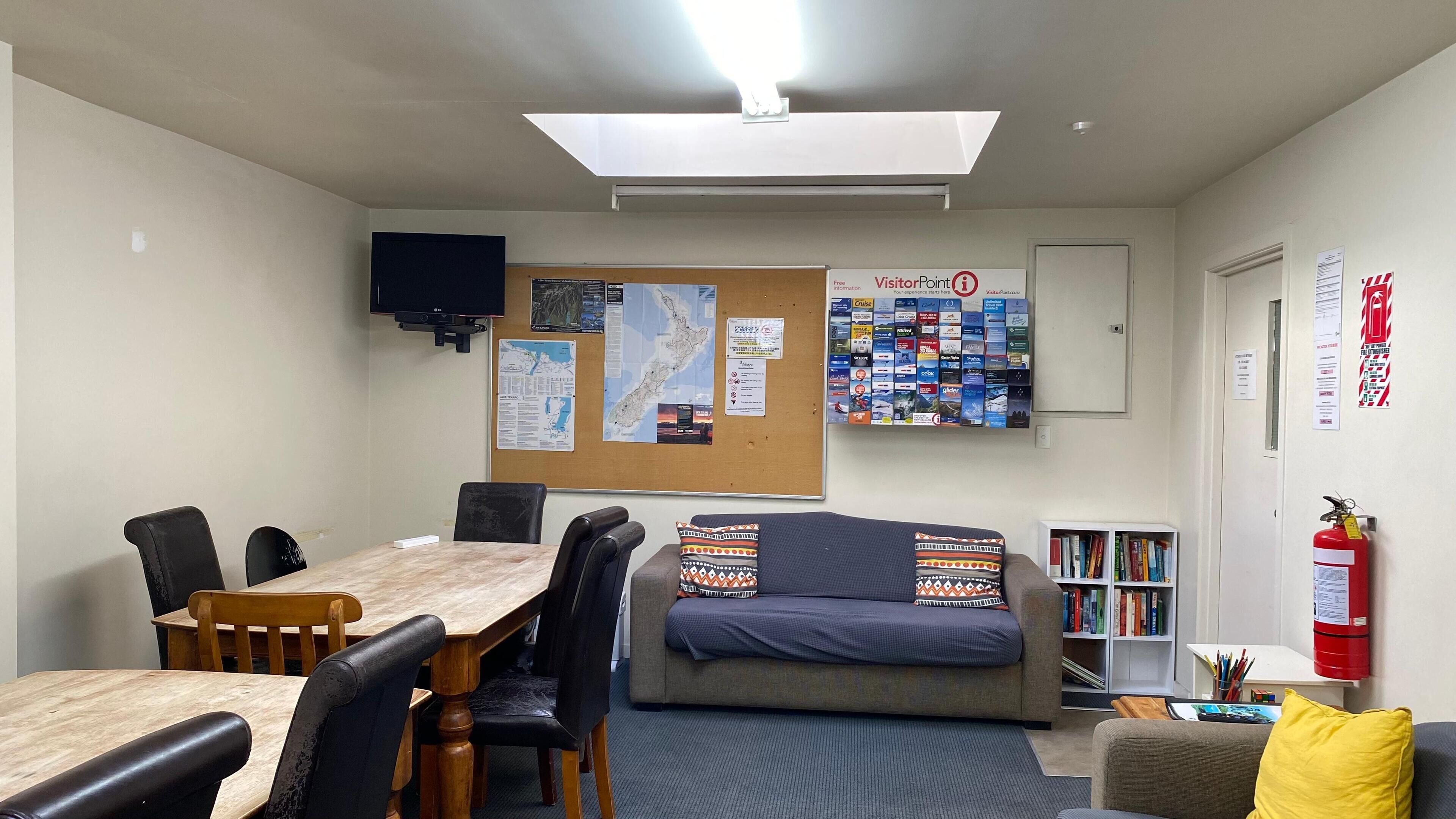 A room with a wooden table, chairs, a sofa with patterned cushions, a bookshelf, a bulletin board with maps and information, and a fire extinguisher.
