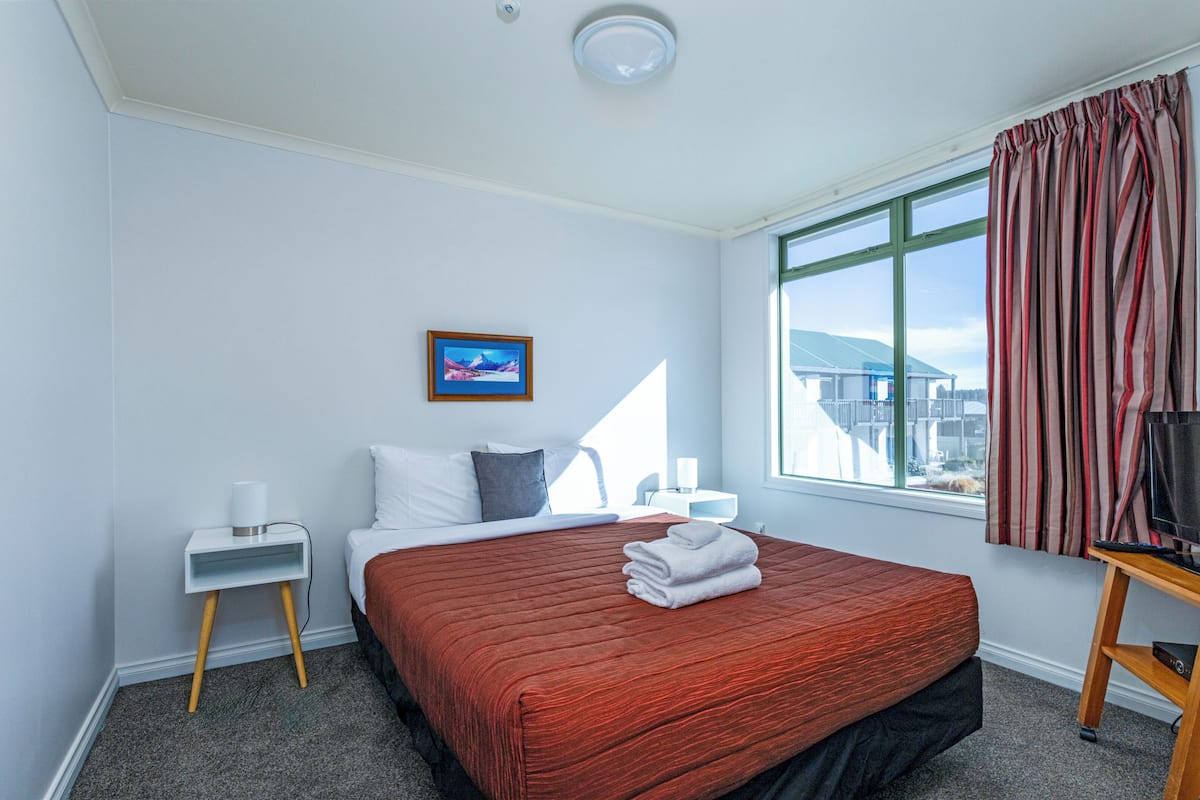 Een hotelkamer met een groot bed, een nachtkastje met lamp, een raam met gordijnen en uitzicht op gebouwen.
