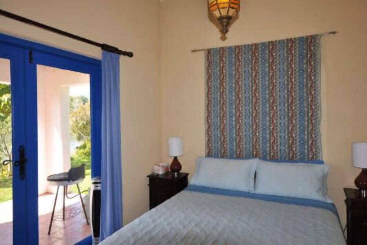 Une chambre à coucher avec un lit, une table de chevet, une lampe, un rideau à motifs et une vue sur l’extérieur par une porte.