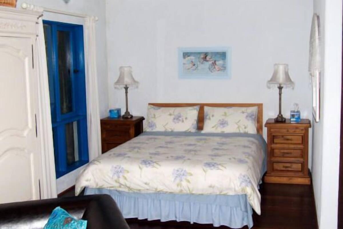Une chambre à coucher avec un lit, deux tables de chevet, une porte bleue, un tableau accroché au mur et un mur blanc.