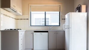 A compact kitchen with white cabinetry, a dishwasher, a refrigerator, and a window with blinds. - Bundalong Holiday Resort (Bundalong)