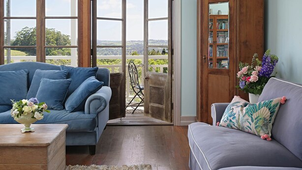 A room with a blue sofa, a grey sofa, a wooden cabinet with books, and a view through open doors. - The Spotted Chook (Montville)