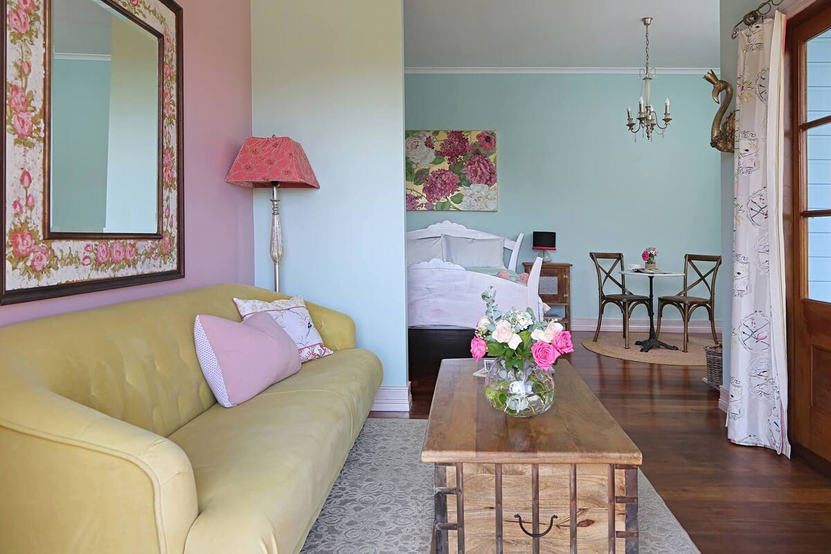 Un salón con sofá amarillo, mesa de centro de madera con un jarrón de flores, un espejo grande con papel tapiz floral, una lámpara y un cuadro en la pared.