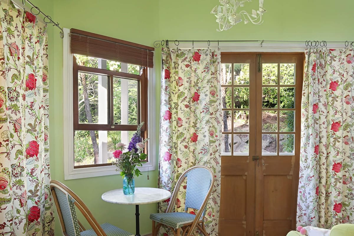 Una habitación con puerta de madera, dos sillas de mimbre, una mesita redonda con un jarrón de flores y cortinas estampadas.