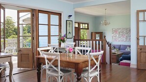 A dining area with a wooden table and white chairs, a staircase, and a view of greenery outside. - The Spotted Chook (Montville)
