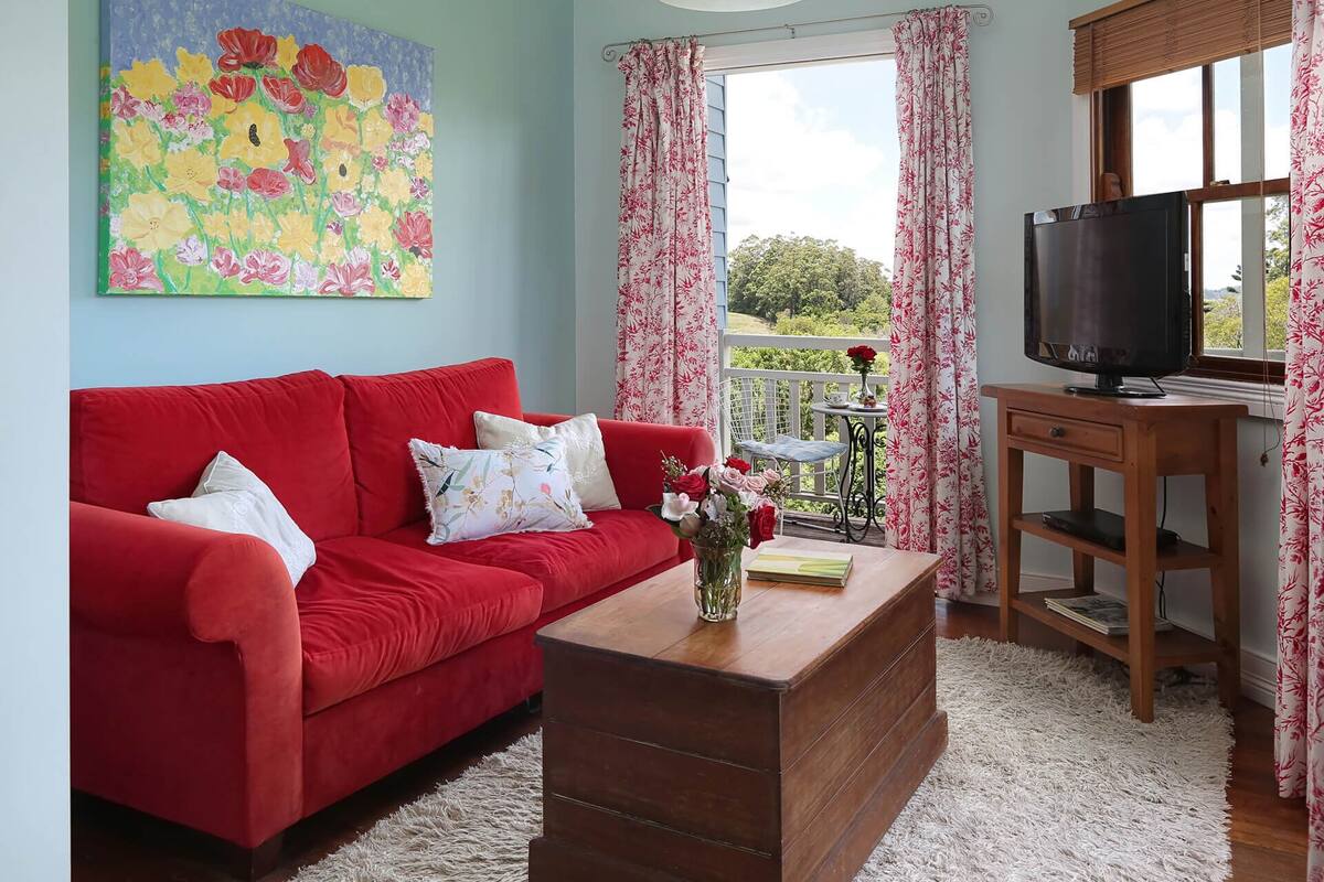 Una sala de estar con un sofá rojo, una mesa de centro de madera con un florero, un televisor sobre un mueble de madera y un cuadro en la pared.