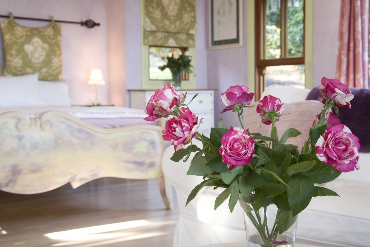 Un dormitorio con una cama, un florero con flores rosadas y una ventana con cortinas.
