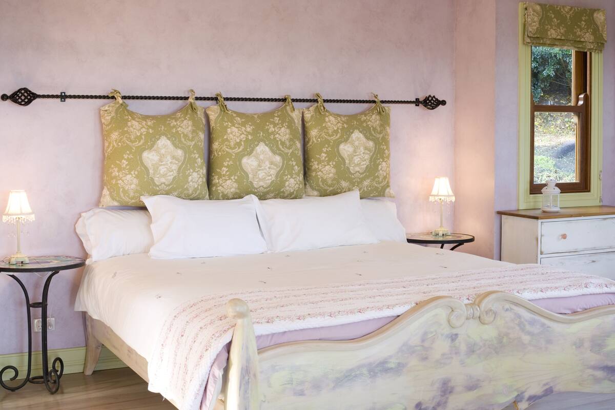Un dormitorio con cama, dos mesitas de noche, una mesilla auxiliar, un ventanal con vistas y una lámpara sobre cada mesita de noche.