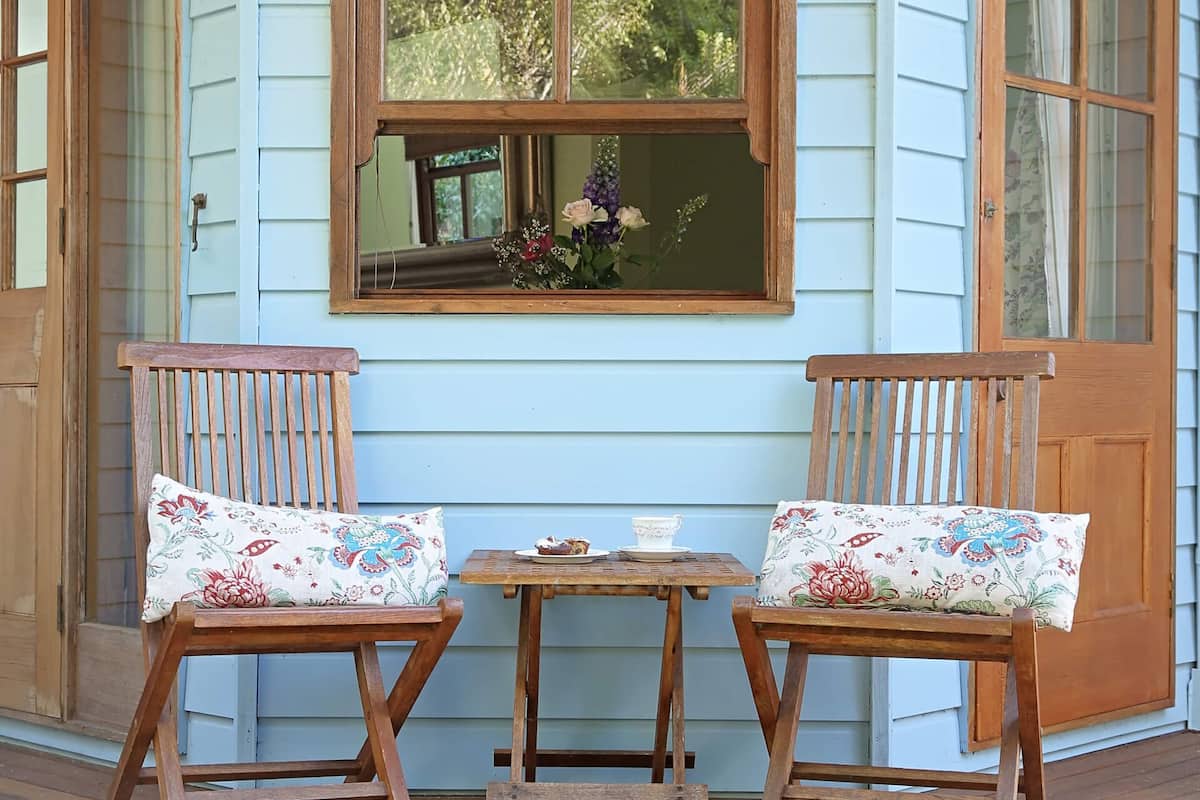 Un porche con dos sillas de madera y una mesita, una ventana decorada con flores y una puerta con un panel de vidrio.