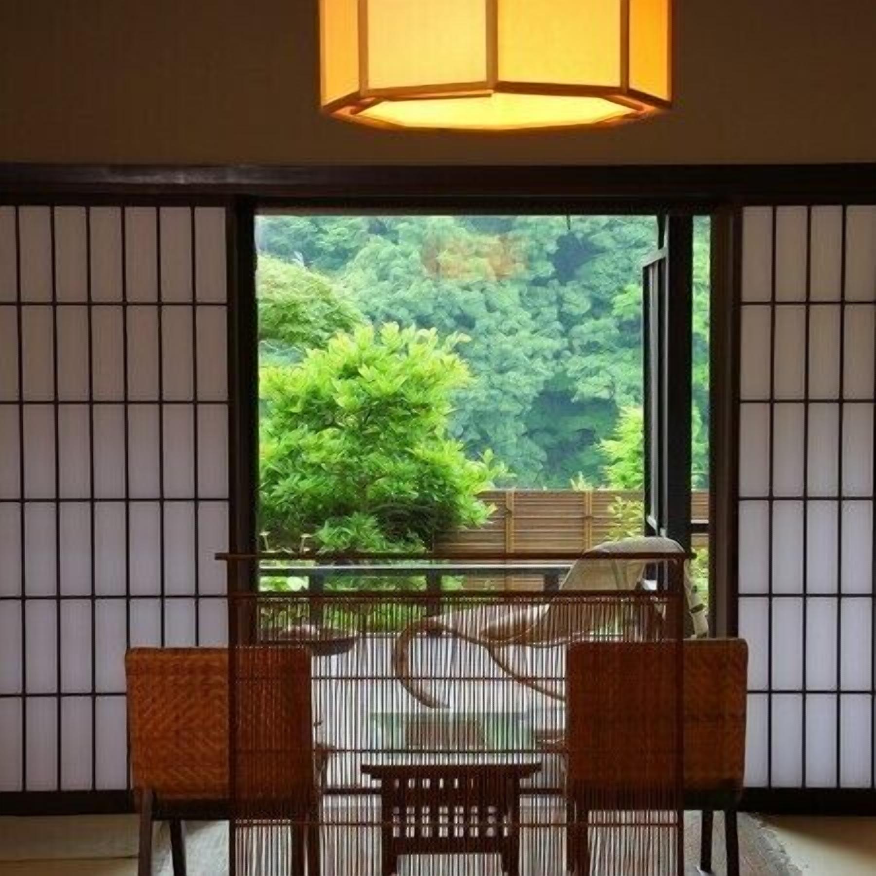 Ein Zimmer mit Tatami-Boden, einem niedrigen Tisch mit zwei Stühlen und Blick auf einen Wald durch eine Schiebetür.