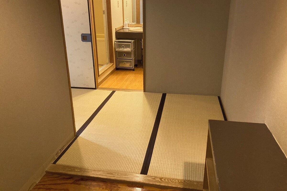 Un couloir étroit au sol recouvert de tatamis, qui mène à une pièce au plancher en bois et dotée d’un petit coin cuisine.