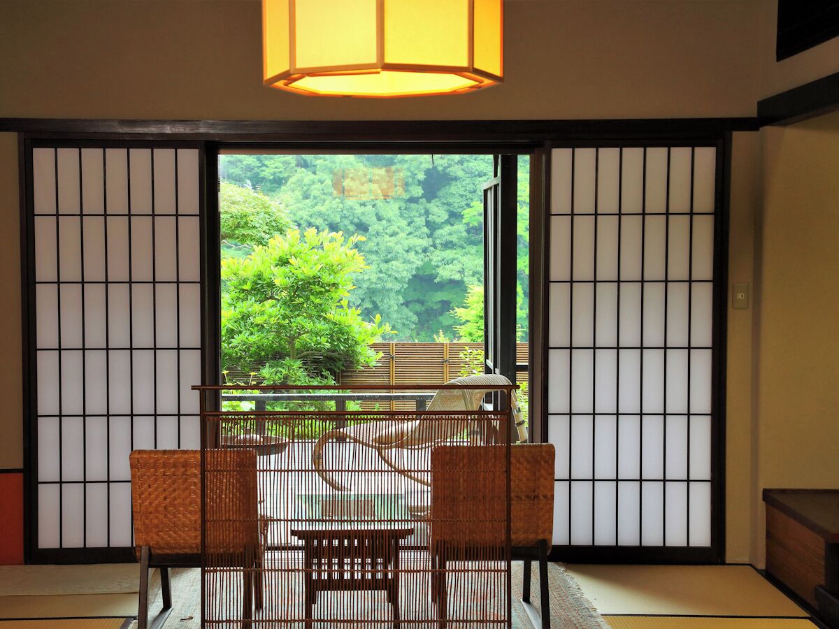 A traditional Japanese room with tatami flooring, a low table with a woven chair, and a view of a lush forest through shoji screens.