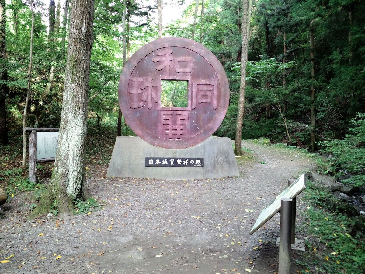 Una placa metálica circular con caracteres japoneses, una base de hormigón y un cartel en una zona boscosa.