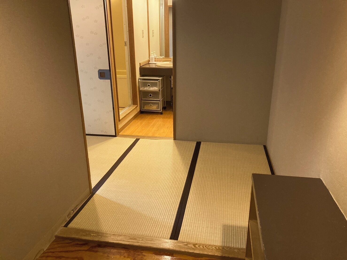 Ein schmaler Gang mit Tatami-Bodenbelag, der zu einem Raum mit Holzboden und einer kleinen Küchenzeile führt.