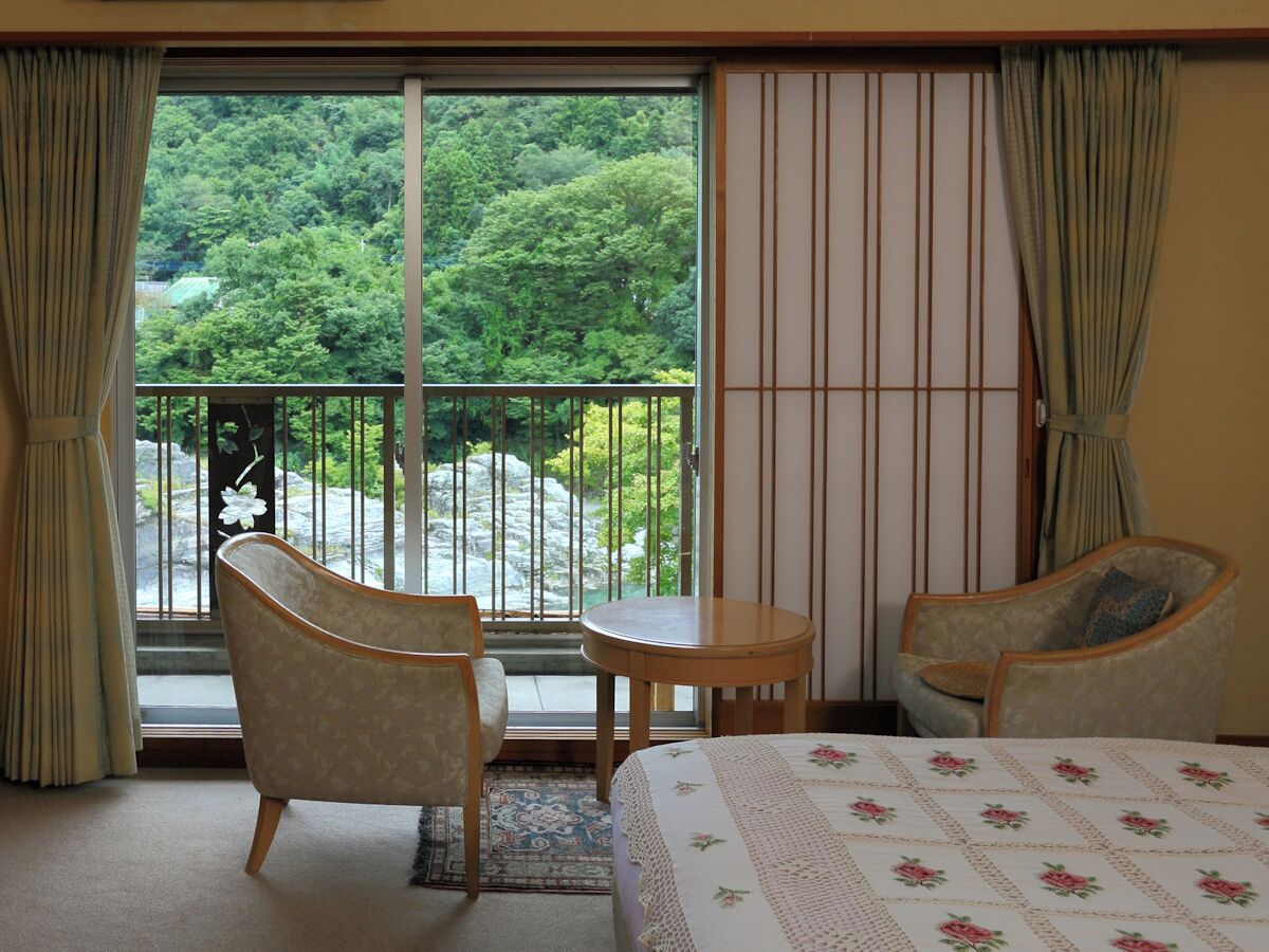 A hotel room with a bed, two armchairs, a small table, and a view of a forest through a sliding door.