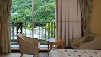A hotel room with a bed, two armchairs, a small table, and a view of a forest through a sliding door.