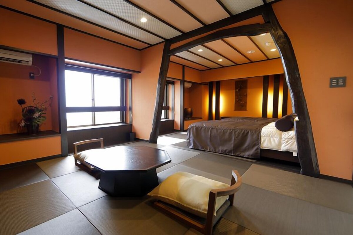 A traditional Japanese-style room with tatami flooring, a low table, a bed, and a window providing natural light.