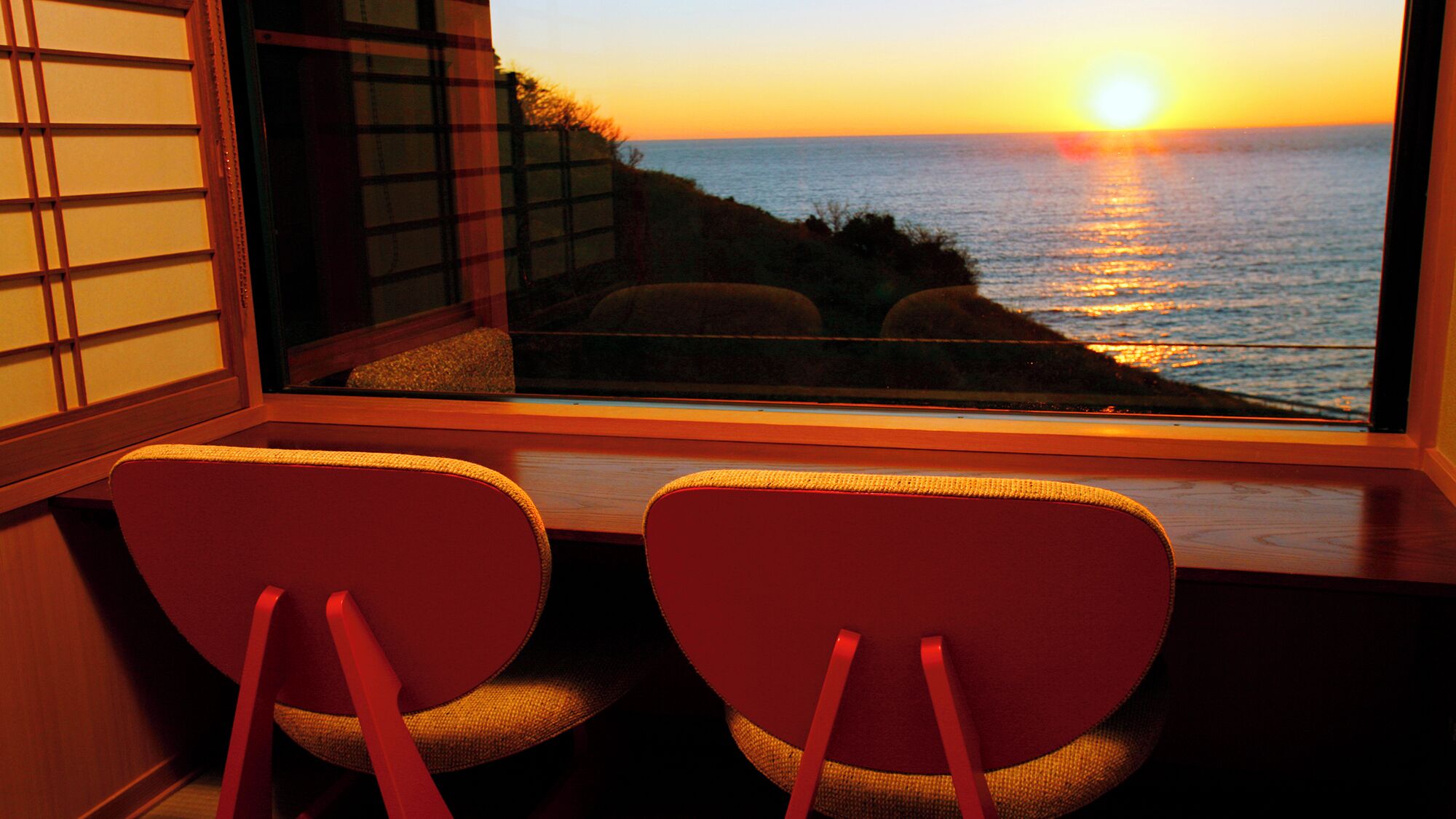 A room with a view of the ocean at sunset, two chairs facing the window, and a traditional Japanese shoji screen.