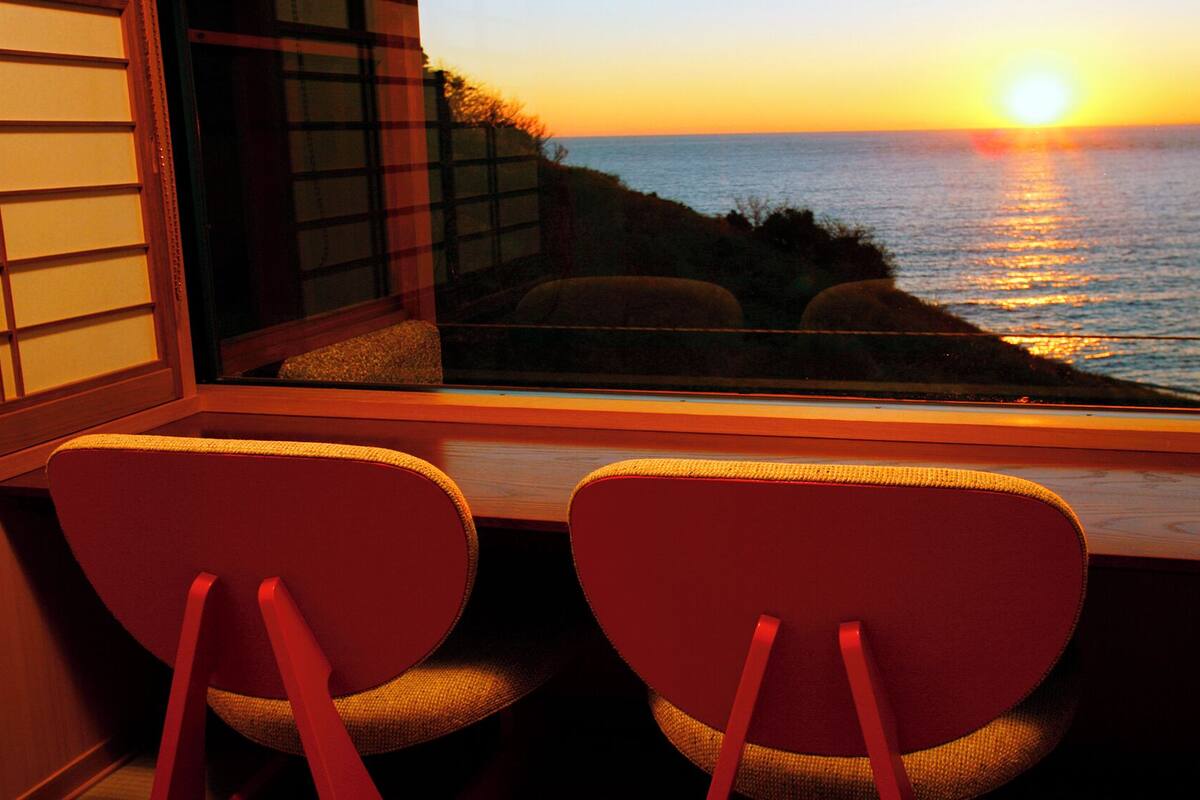 Ein Zimmer mit Meerblick bei Sonnenuntergang, zwei Sessel zum Fenster hin und ein traditioneller japanischer Shoji-Bildschirm.