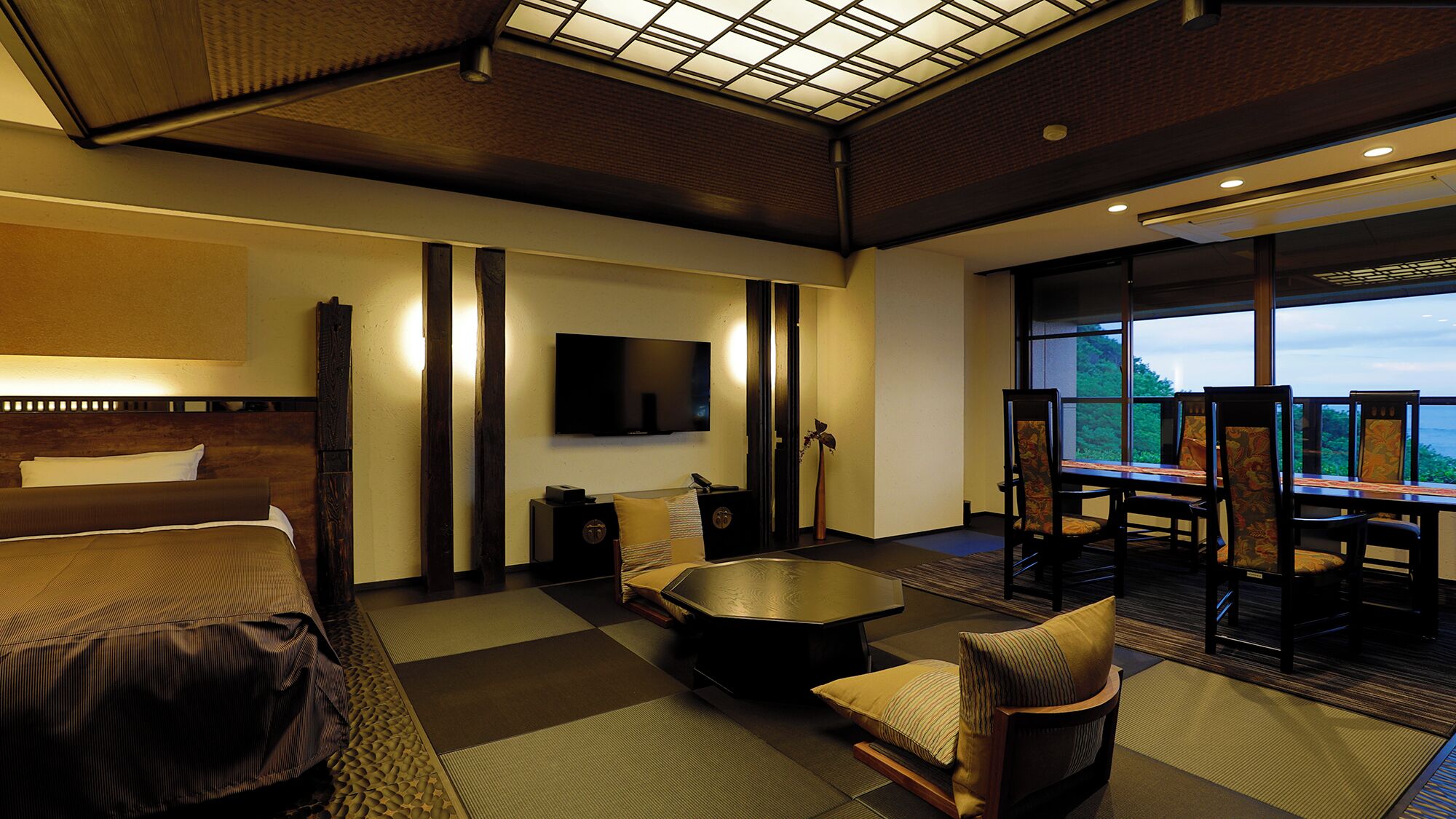 A hotel room with a bed, a TV, a dining area with chairs and a table, and a view through a glass wall.