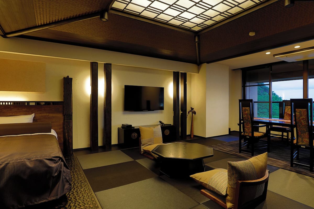 Ein Hotelzimmer mit Bett, Fernseher, Essecke mit Stühlen und Tisch sowie Blick durch eine Glaswand.