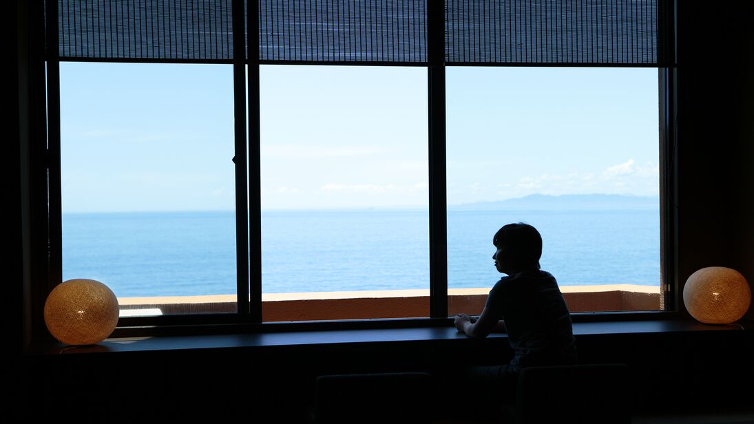 Una habitación con un ventanal grande que da a una playa y al océano, un niño sentado junto a la ventana, y dos lámparas esféricas en el alféizar.