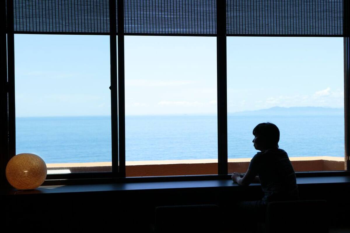 Ein Zimmer mit großem Fenster mit Blick auf Strand und Meer, ein Kind sitzt am Fenster, und zwei runde Lampen stehen auf der Fensterbank.