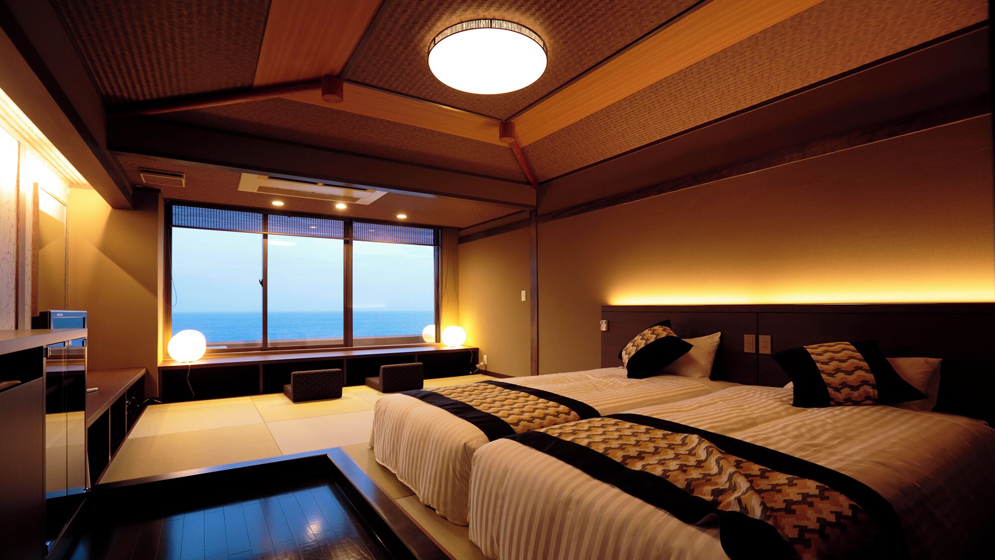 A hotel room with a large window, a bed, a bedside table, and a small desk with a lamp.
