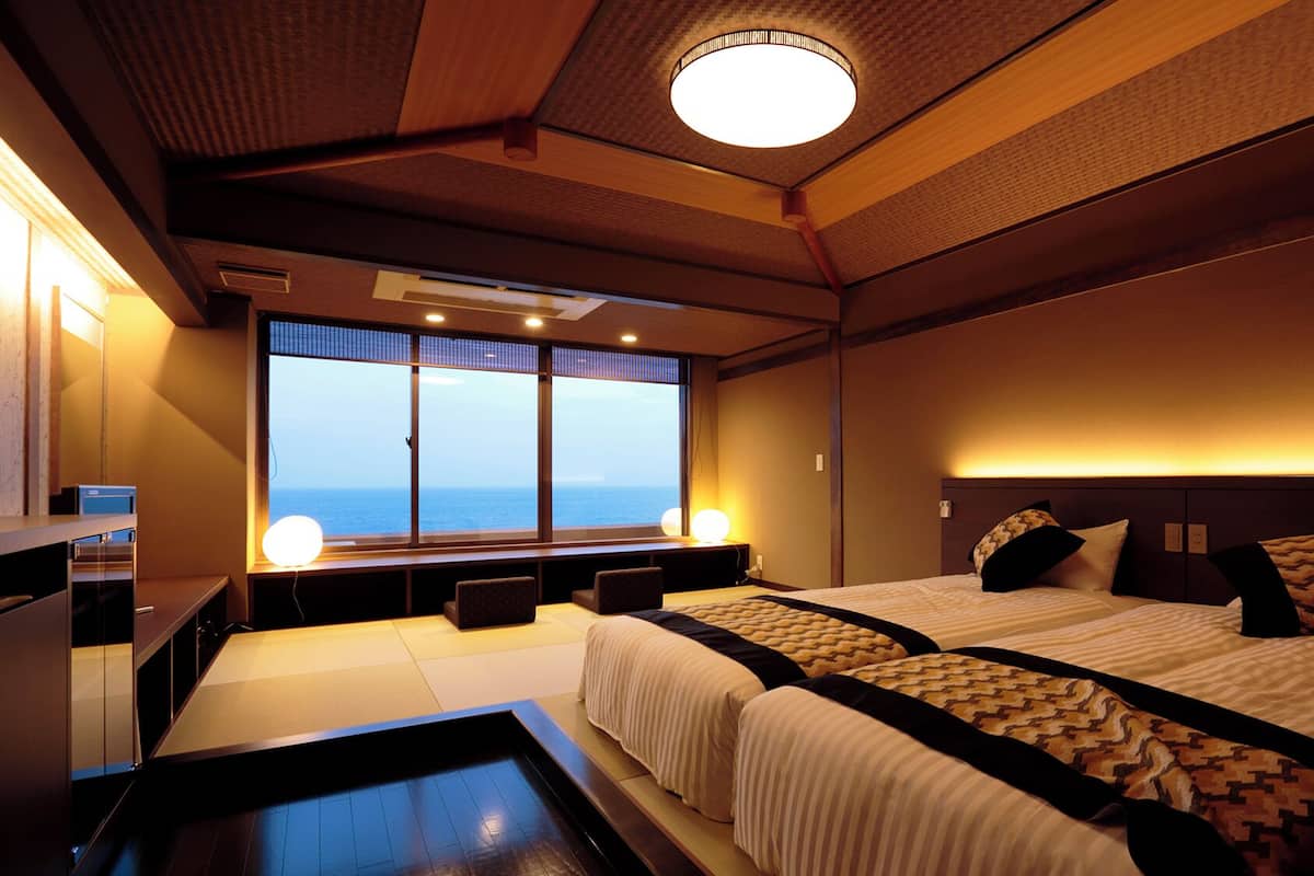 Ein Hotelzimmer mit großem Fenster, einem Bett, einem Nachttisch und einem kleinen Schreibtisch mit Lampe.