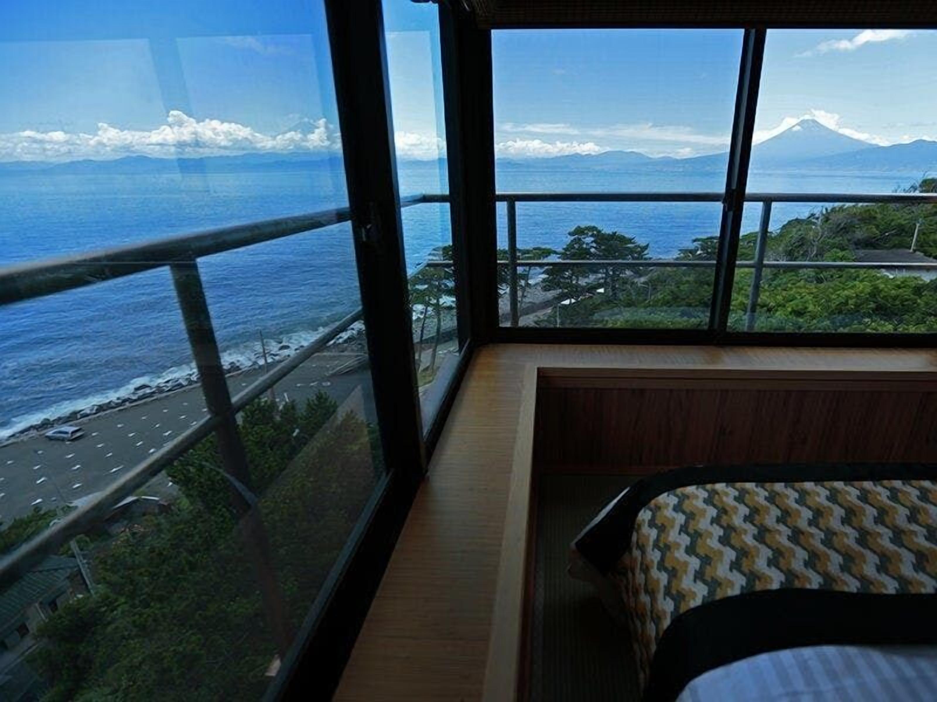 A room with a large window overlooking a coastal road and a mountain.