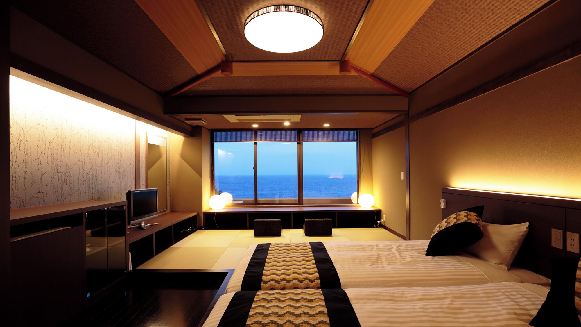 A hotel room with a large bed, a TV, a window with a view of the sea, and a ceiling light.