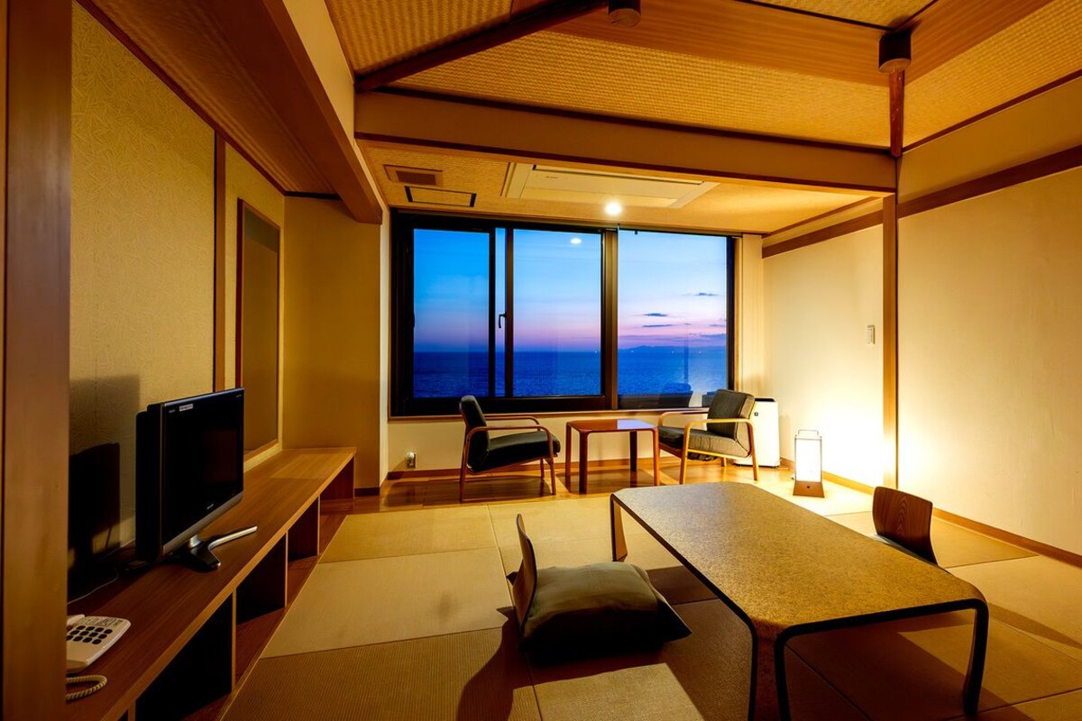A room with tatami flooring, a television, a desk, and a view of the sunset through large windows.