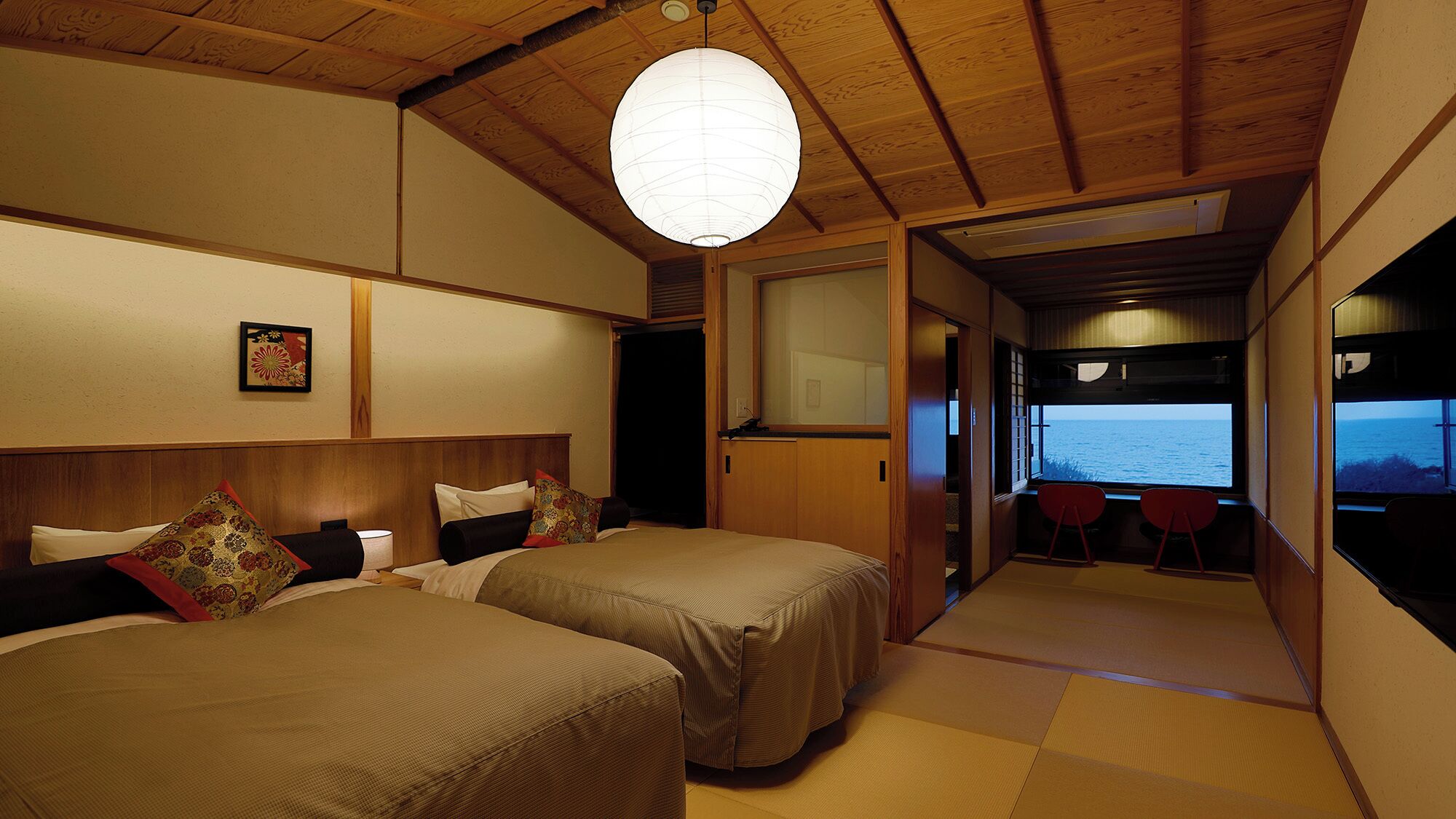 A room with two beds, a hanging paper lantern, a view of the sea, and tatami flooring.