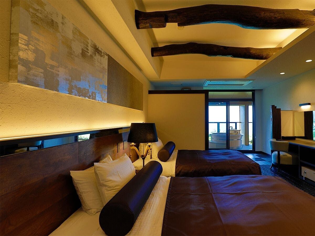 A hotel room with a large bed, a desk, a chair, a lamp, and a large window.