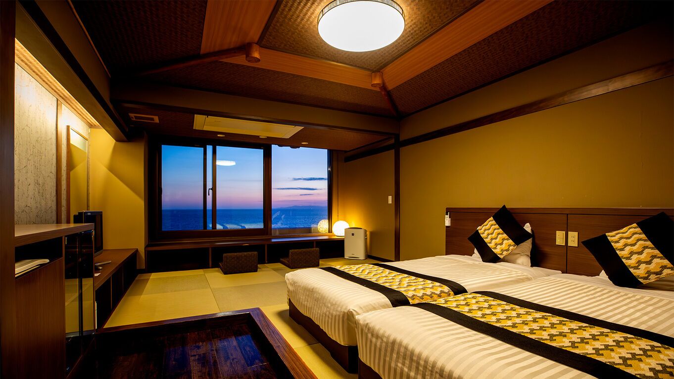 A hotel room with two beds, a TV, a desk, and a view of the ocean.
