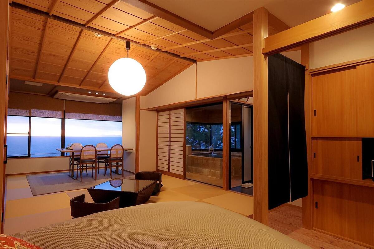 A room with a view of the ocean, a dining area with chairs, and a sliding door leading to a balcony.