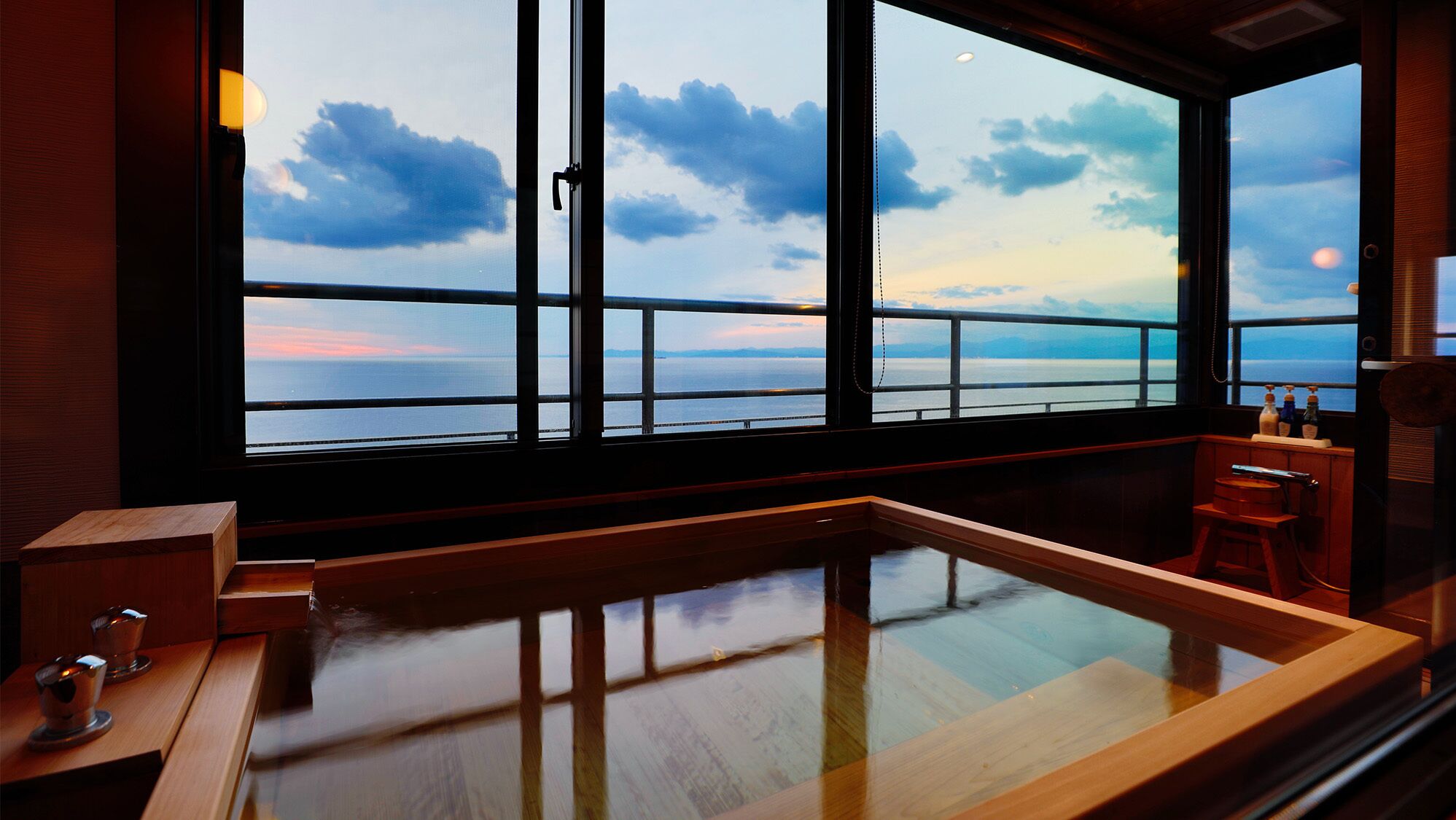 A room with a large window overlooking a body of water, a wooden bench, and a small table with bottles.