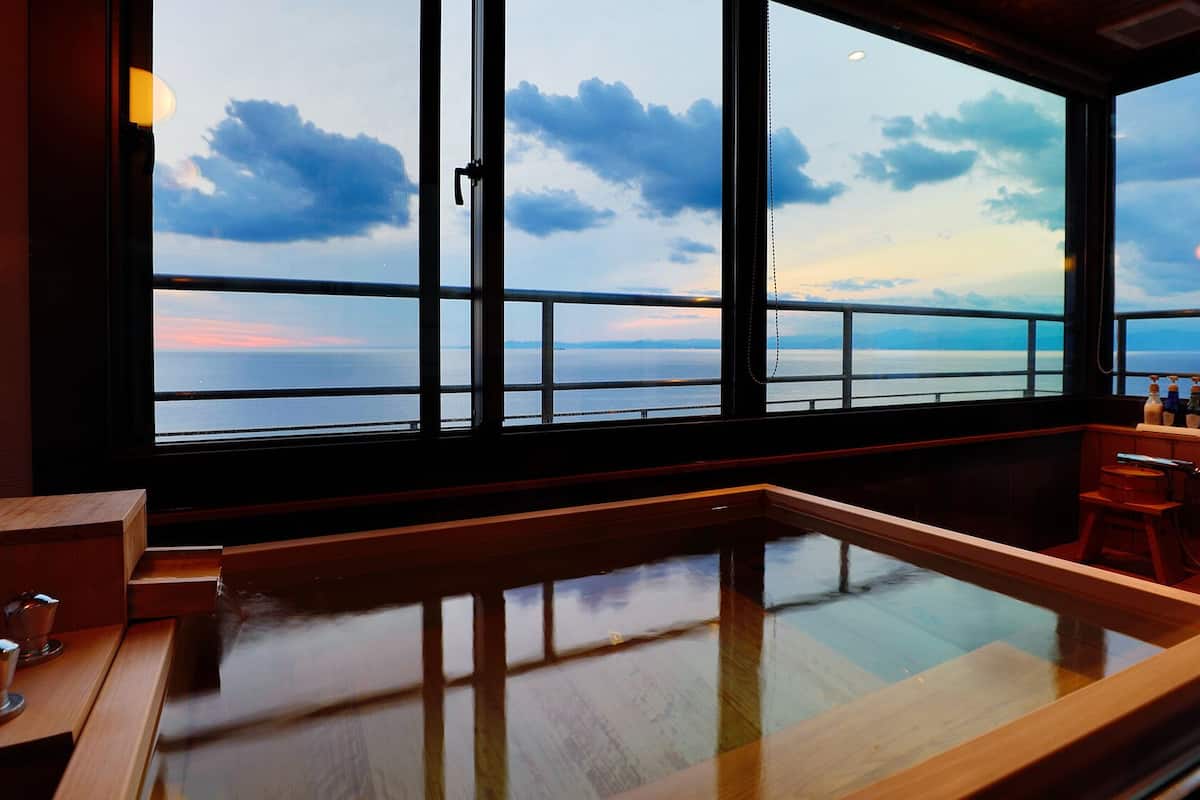 Ein Zimmer mit großem Fenster und Blick auf ein Gewässer, einer Holzbank und einem kleinen Tisch mit Flaschen.