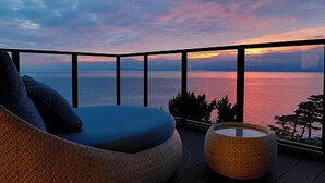 A balcony with a view of the sea during sunset, a wicker sofa, and a round wicker ottoman. - Umi No Hotel Isaba (Numazu)