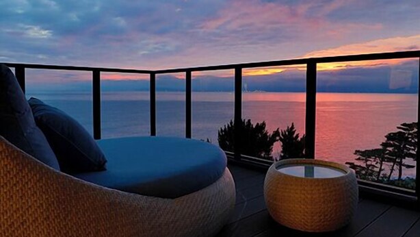 A balcony with a view of the sea during sunset, a wicker sofa, and a round wicker ottoman.