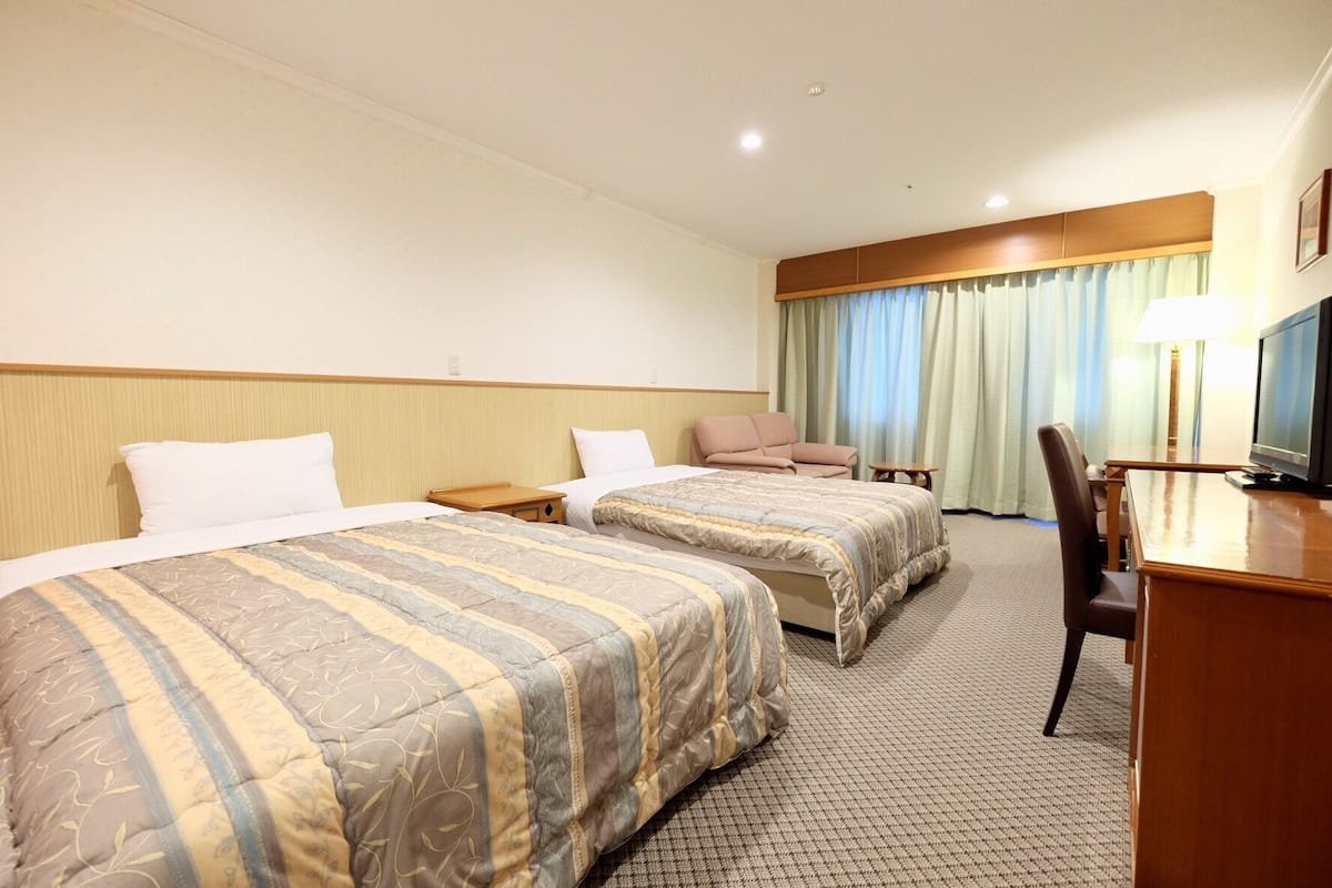 A hotel room with two beds, a desk with a television, a chair, and a window with curtains.