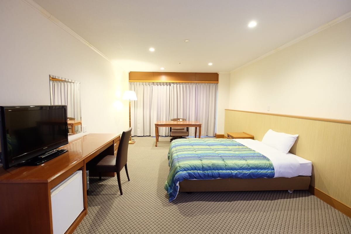 A hotel room with a bed, a desk with a TV, a chair, a table, and a lamp.