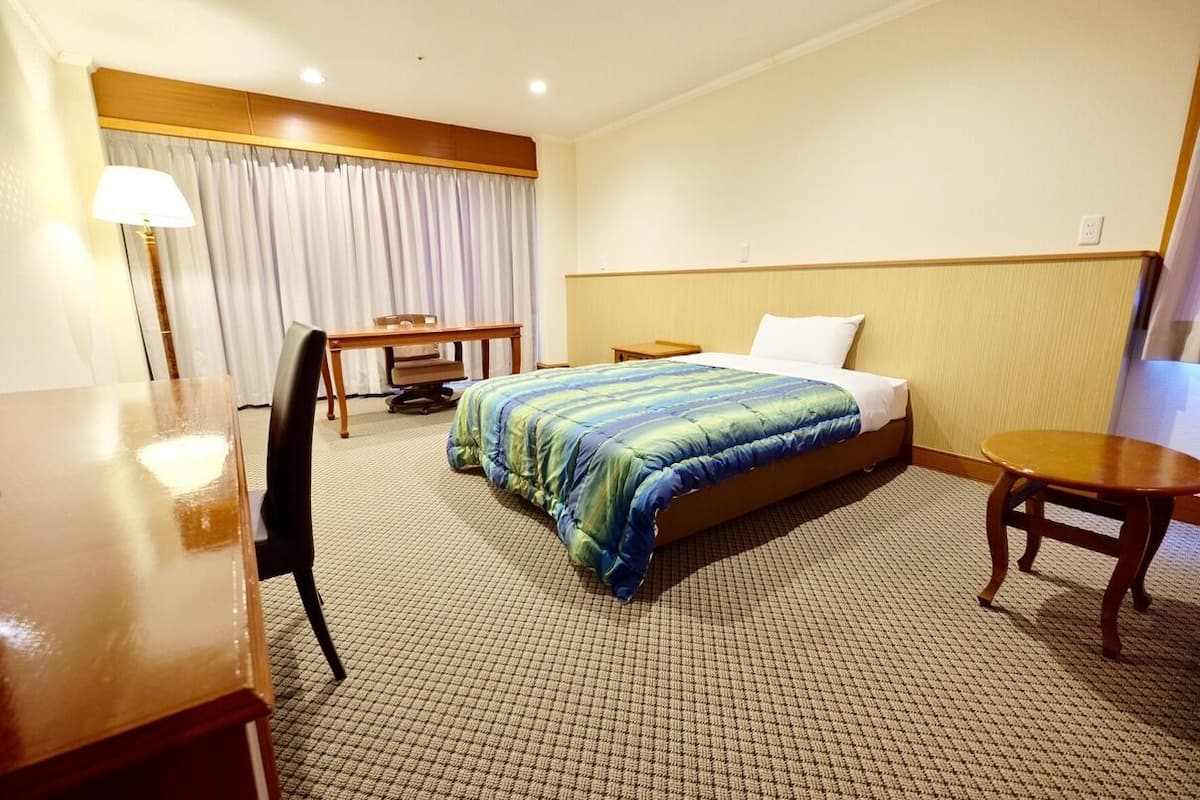 A hotel room with a bed, a desk, a chair, a small table, and a lamp.