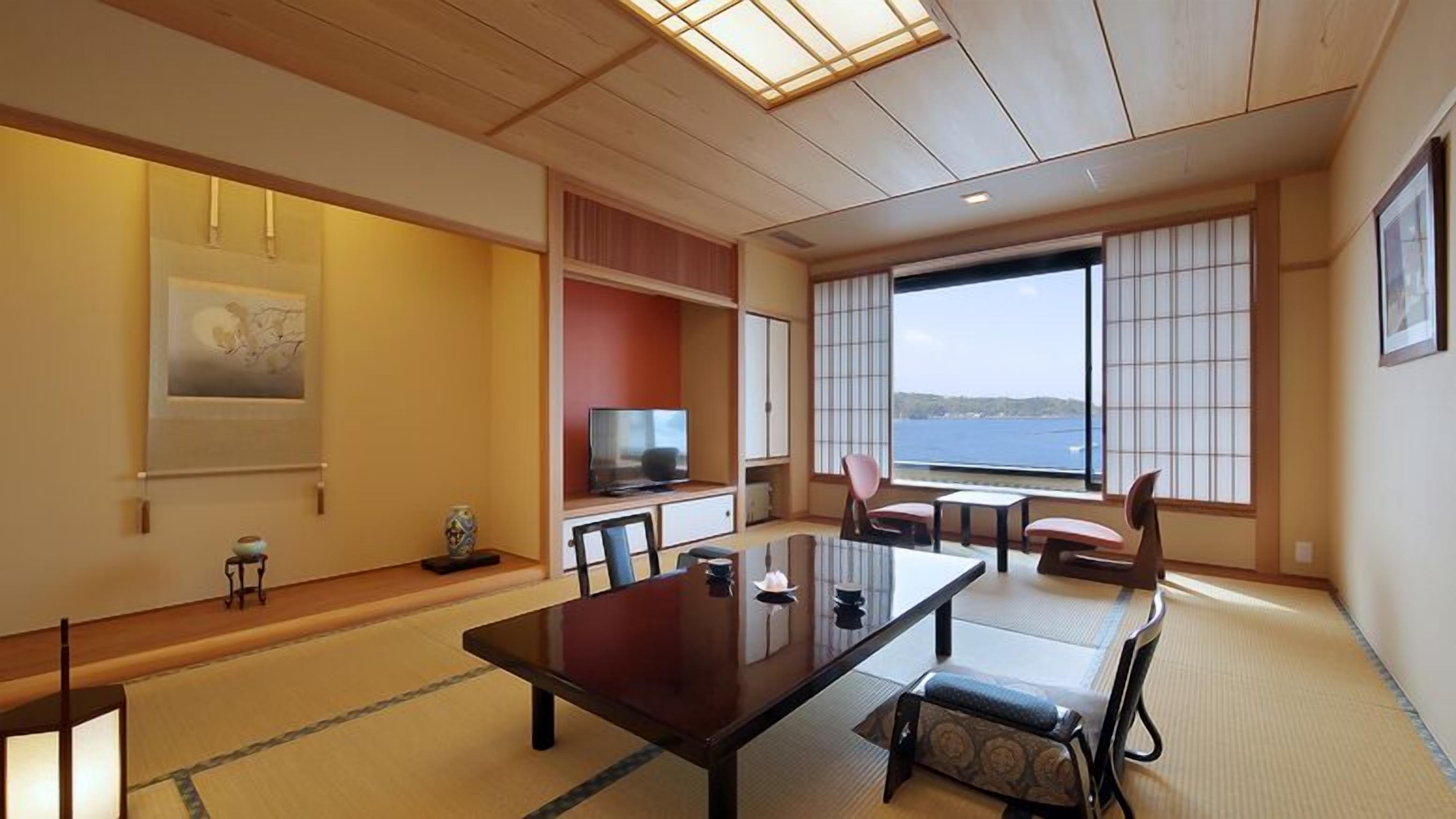 A traditional Japanese-style room with tatami flooring, a low table, chairs, a television, and a large window with a view of the sea.