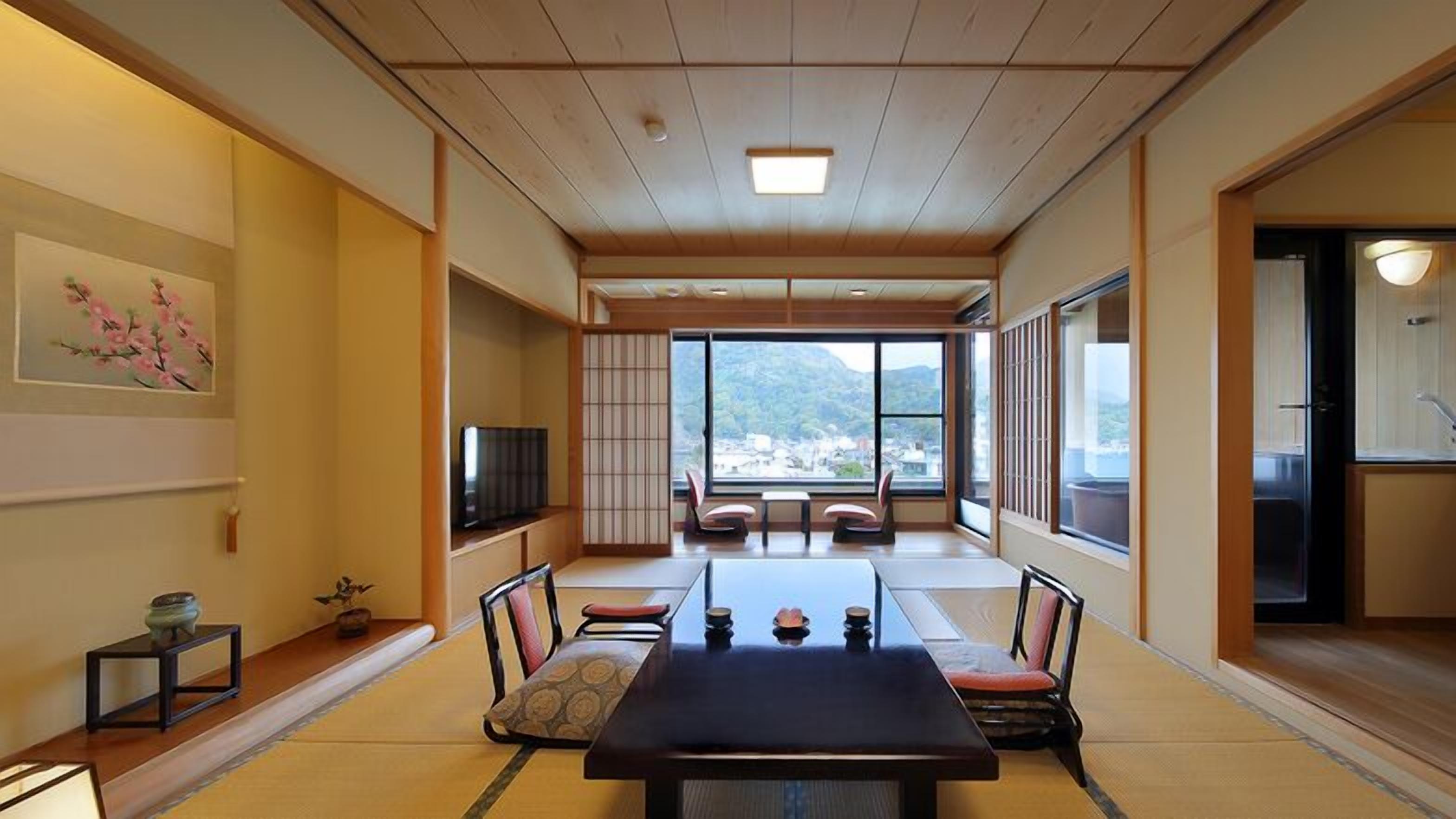 A traditional Japanese-style room with tatami flooring, a low table with cushions, a television, and a view of a mountainous landscape.