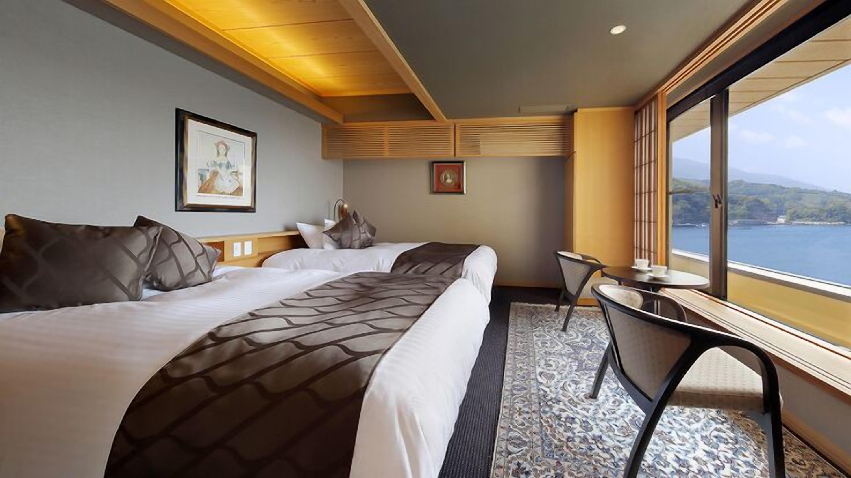 A hotel room with two beds, a small table, a chair, a window with a view of the sea, and artwork on the walls.