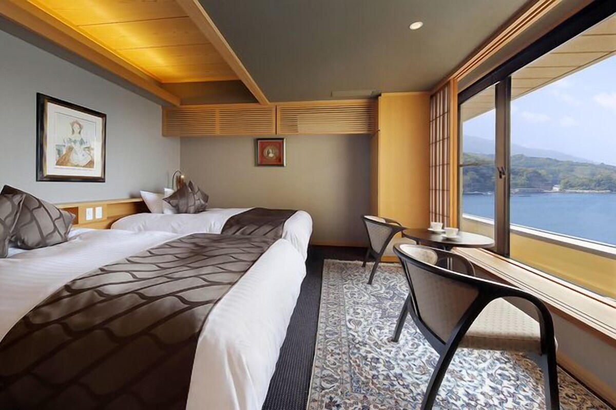 A hotel room with two beds, a small table, a chair, a window with a view of the sea, and artwork on the walls.