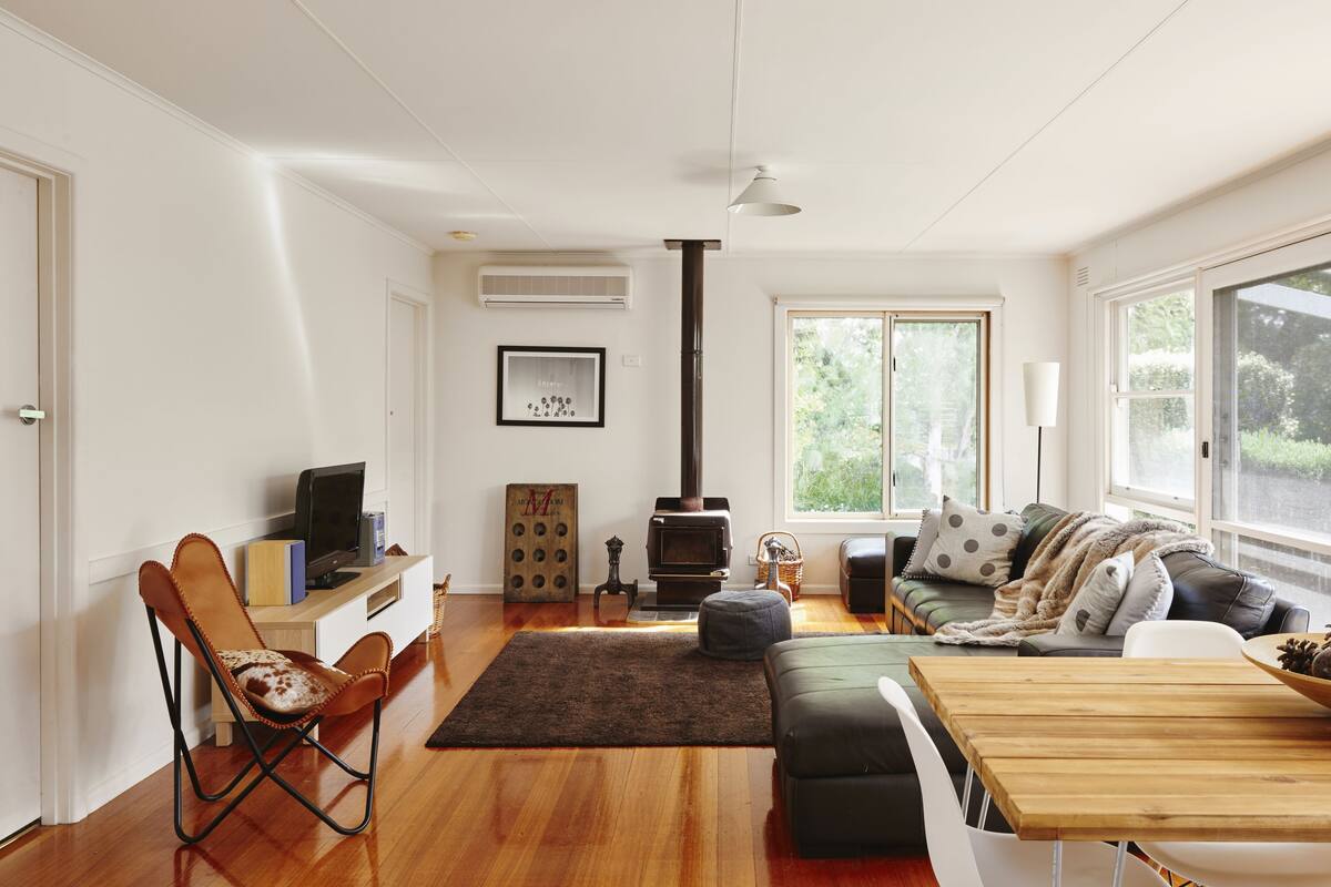 A spacious living room with a wooden floor, a sofa set, a television, a fireplace, and large windows offering a view of greenery.