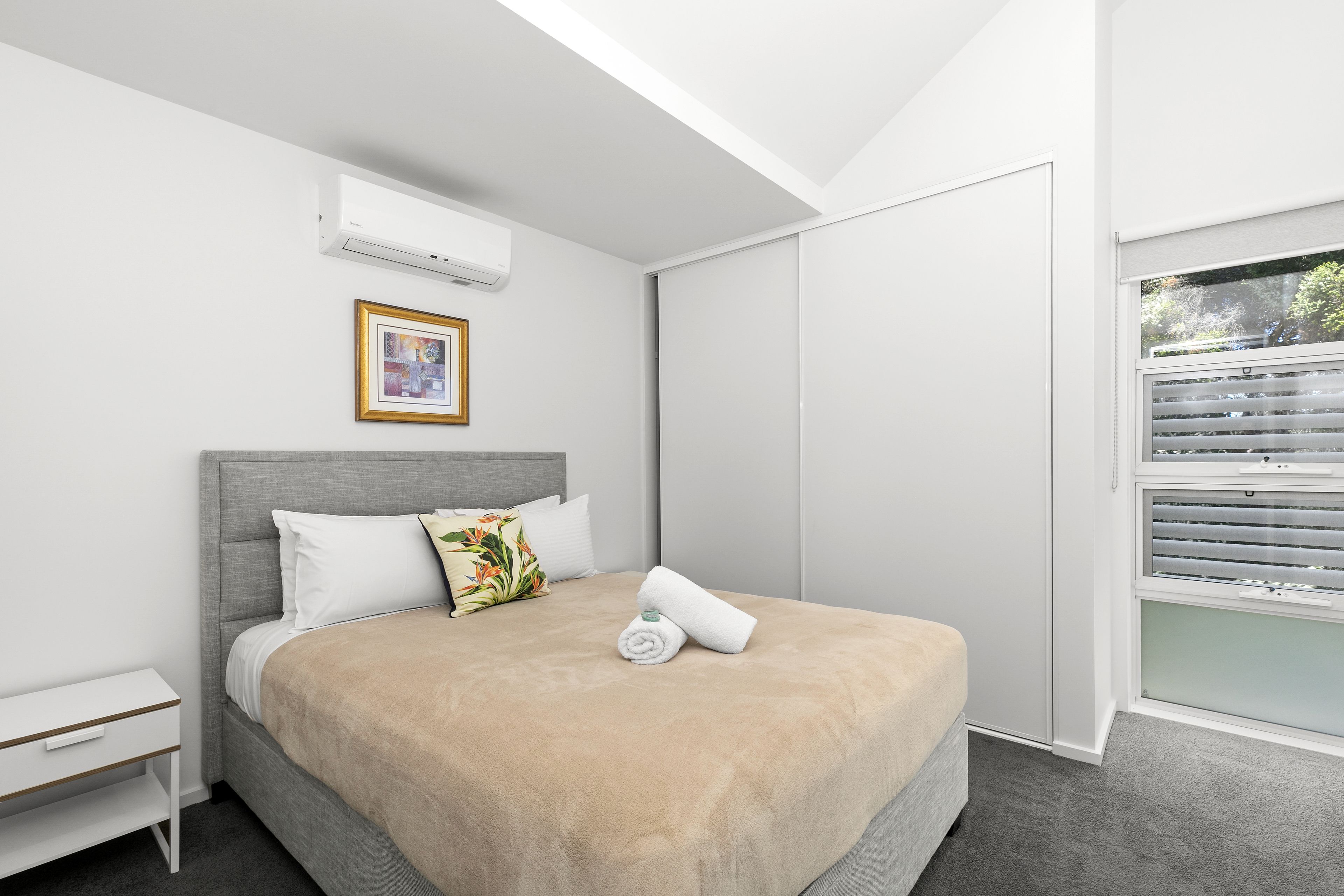 A modern bedroom with a bed, a nightstand, a framed picture, and a window with blinds.