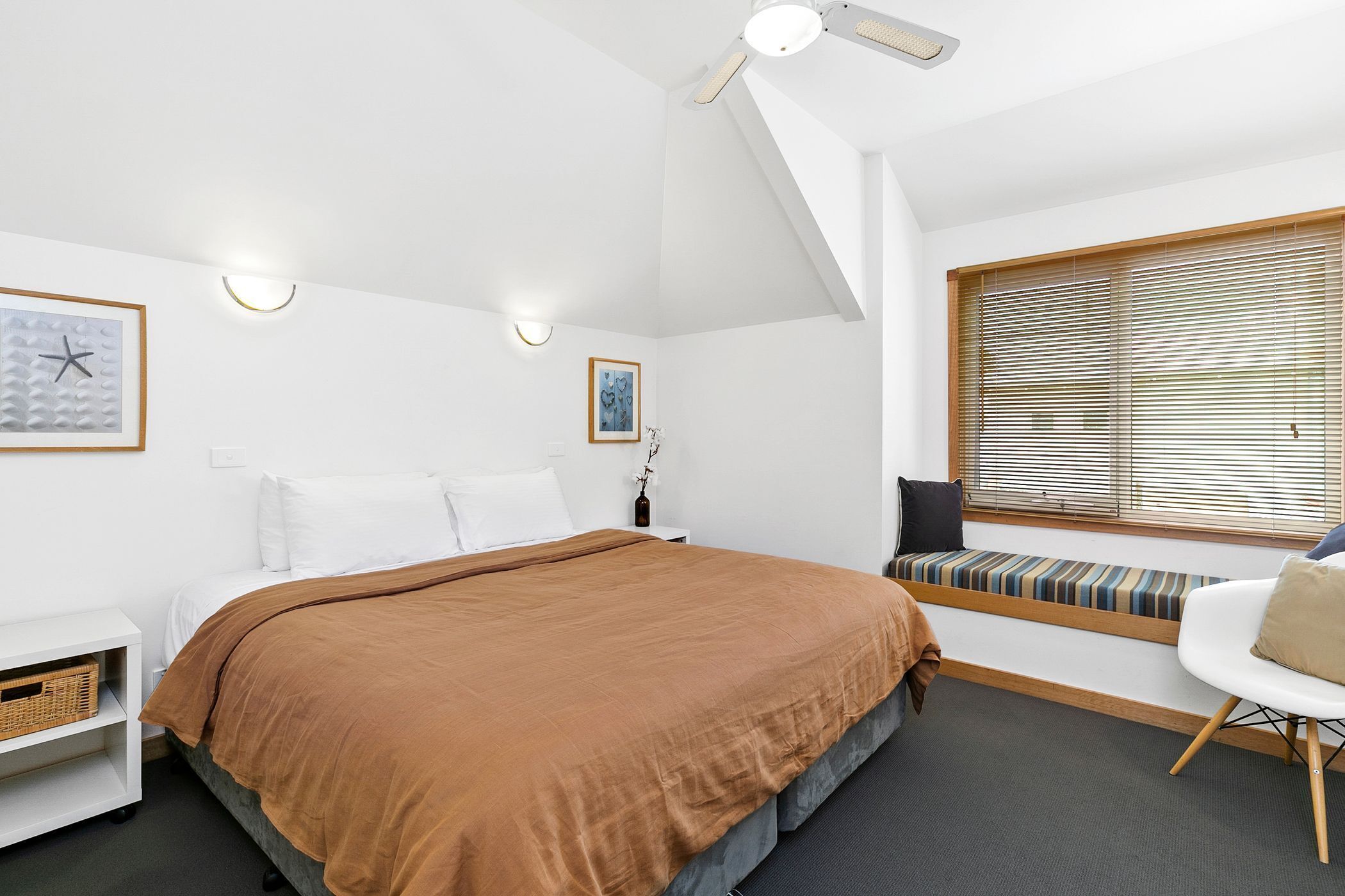 A bedroom with a large bed, a bedside table, a chair, a window with blinds, and wall-mounted lights.